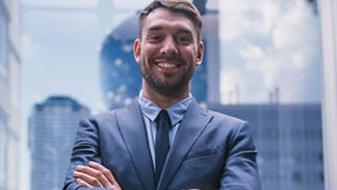 Homem de negócios sorridente, de fato azul e gravata, com os braços cruzados, posando em frente a edifícios corporativos de vidro.