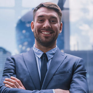 A smiling businessman in a blue suit and tie, with his arms crossed, posing in front of glass corporate buildings.
