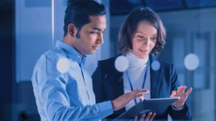 Dois profissionais, um homem e uma mulher, a analisar informações num tablet num ambiente de escritório moderno com iluminação azulada.