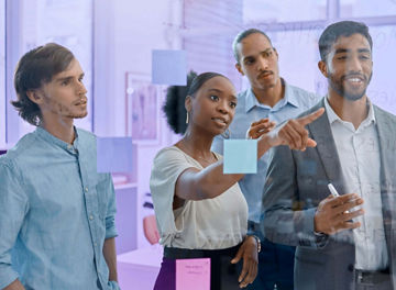 A group of five professionals collaborating during a work session, using sticky notes and a glass whiteboard in a modern office.