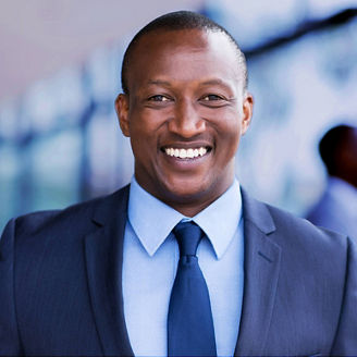 A smiling businessman in a blue suit and tie, with his arms crossed, posing in front of glass corporate buildings.