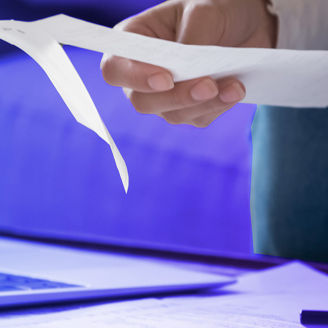 A hand is typing something on a calculator and holding a piece of paper in front of a laptop.