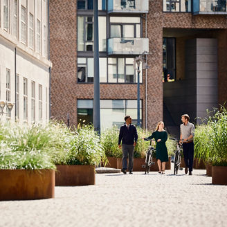 Three people walking along a cobblestone path in a modern urban area with large green planters and bicycles
