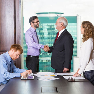 Smiling senior and young businessmen standing at head of table and shaking hands. Businesswoman is standing at table and looking at them. Another businessman is sitting at table and making notes. 