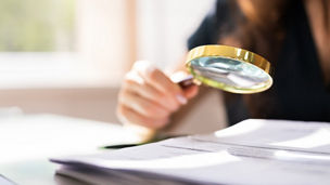 Someone is holding a magnifying glass over papers next to a laptop.
