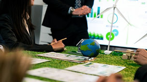 People working around a table with sustainability-related materials, including a small globe, wind turbine model, and documents featuring graphs and charts.