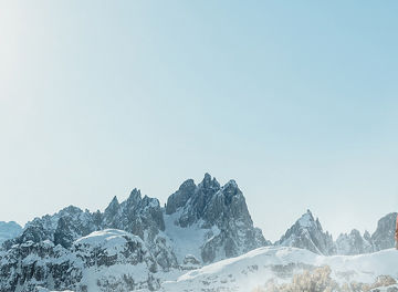 Das Bild zeigt eine Person in Outdoor-Kleidung mit einem Rucksack, die auf eine schneebedeckte Berglandschaft blickt. Die Berge sind majestätisch und von Schnee bedeckt, während der Himmel klar und blau ist, was eine ruhige und erhabene Atmosphäre schafft. Die Szene vermittelt ein Gefühl von Abenteuer und Naturverbundenheit.