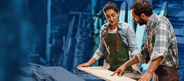 Zwei Personen stehen in einer Werkstatt und arbeiten gemeinsam an einem Holzbrett. Die Umgebung ist unscharf, während die Personen und das Brett im Vordergrund klar zu erkennen sind.