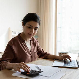 Household budget. Young indian female sit at table calculate utility payments before make money transfer in online bank app on laptop. Focused millennial business woman pay taxes plan personal savings