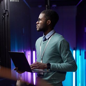 Side view portrait of African American data engineer holding laptop while working with supercomputer in server room lit by blue light, copy space