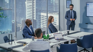 Konferenzraum in einem Hochhaus: Vier Personen in Businesskleidung, ein Mann präsentiert Folien auf einem Bildschirm, Laptops und Notizen auf dem Tisch.