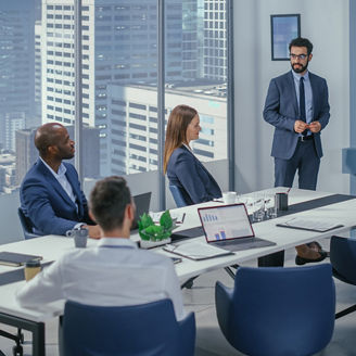Konferenzraum in einem Hochhaus: Vier Personen in Businesskleidung, ein Mann präsentiert Folien auf einem Bildschirm, Laptops und Notizen auf dem Tisch.