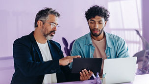 Professional men are engaged in a productive discussion in a brightly lit office. They use a tablet and a laptop to discuss and brainstorm business matters, highlighting teamwork and collaboration.
