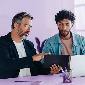 Professional men are engaged in a productive discussion in a brightly lit office. They use a tablet and a laptop to discuss and brainstorm business matters, highlighting teamwork and collaboration.