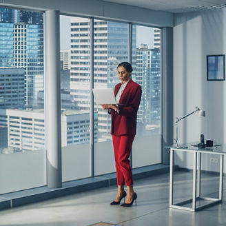 Successful Caucasian Businesswoman Using Laptop While Standing in Office Looking out of Window on Big City. Perfect Confident Female Digital Entrepreneur Developing e-Commerce Software Strategy.
