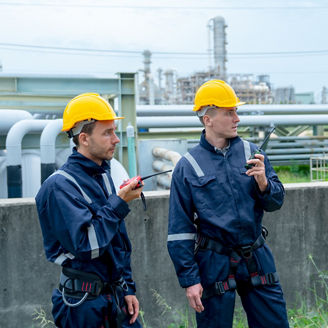 Two petrochemical or gas factory workers discuss about work with look to left side and stand in front of pipeline of factory.