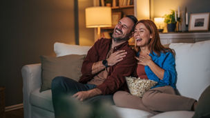 A husband and wife are watching a movie at night and eating popcorn in the living room.