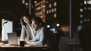 Two focused young business colleagues working overtime together over a laptop at a desk late in the evening