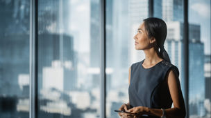 Successful Businesswoman in Stylish Dress Standing in Modern Office, Using Smartphone, Looking out of the Window on Big City with Skyscrapers. Confident Female CEO Working on Financial Projects.