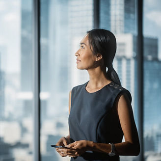 Successful Businesswoman in Stylish Dress Standing in Modern Office, Using Smartphone, Looking out of the Window on Big City with Skyscrapers. Confident Female CEO Working on Financial Projects.