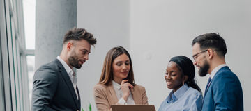 Four businesspeople are standing together in a modern office, collaborating and reviewing information on a clipboard, engaging in productive discussion