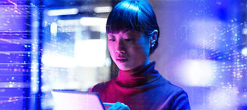 A woman olding and touching a tablet with both hands, illuminated by blue and pink lighting, with abstract digital light patterns and blurred structures in the background