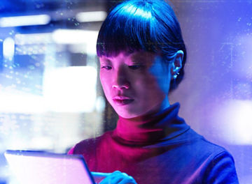 A woman olding and touching a tablet with both hands, illuminated by blue and pink lighting, with abstract digital light patterns and blurred structures in the background