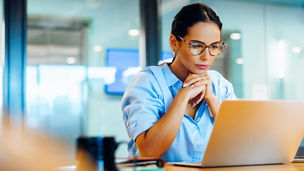 Focused businesswoman wearing glasses engrossed in work at a laptop computer in an office environment.
