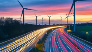Autoestrada ao entardecer com trilhos de luz e aerogeradores eólicos sob um céu colorido.