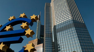 big-blue-euro-sign-with-golden-stars-in-front-of-tall-building.jpg