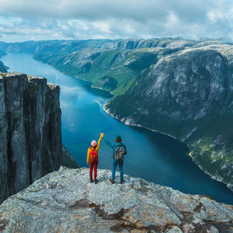 Fotografía de dos excursionistas sobre un acantilado rocoso con vista panorámica a un fiordo rodeado de montañas escarpadas y agua azul bajo un cielo nublado