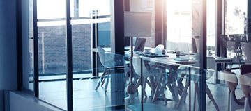 Das Bild zeigt einen modernen Konferenzraum mit einem großen Tisch und mehreren Stühlen. Der Raum ist durch große Glasfenster und -wände gekennzeichnet, die viel Licht hereinlassen und eine helle, offene Atmosphäre schaffen. Auf dem Tisch befinden sich einige Notizbücher und Tassen. Die Einrichtung ist minimalistisch und in neutralen Farben gehalten.