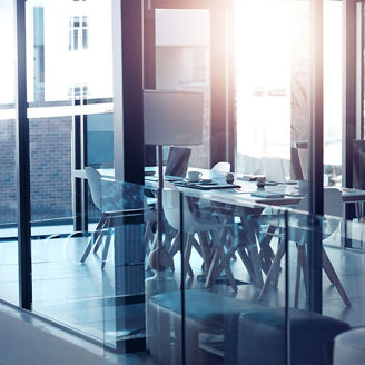 Das Bild zeigt einen modernen Konferenzraum mit einem großen Tisch und mehreren Stühlen. Der Raum ist durch große Glasfenster und -wände gekennzeichnet, die viel Licht hereinlassen und eine helle, offene Atmosphäre schaffen. Auf dem Tisch befinden sich einige Notizbücher und Tassen. Die Einrichtung ist minimalistisch und in neutralen Farben gehalten.