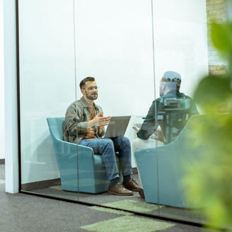 Two professionals engage in a conversation while seated comfortably in an office lounge, surrounded by greenery and contemporary design elements