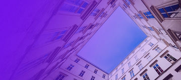 Ein Blick von unten in einen quadratischen Innenhof, umgeben von hohen Gebäuden mit weißen Fassaden und zahlreichen Fenstern. Der Himmel ist klar und blau, sichtbar durch die offene Mitte des Hofes. Der linke Teil des Bildes ist mit einem violetten Farbverlauf überlagert, der dem Bild eine moderne und künstlerische Note verleiht.