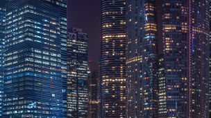 Cityscape view of modern office business buildings in a major financial district. The buildings have glass facades and are illuminated at night, showing lit windows.