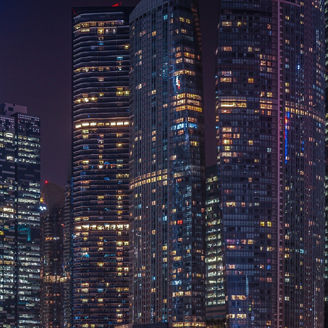 Cityscape view of modern office business buildings in a major financial district. The buildings have glass facades and are illuminated at night, showing lit windows.