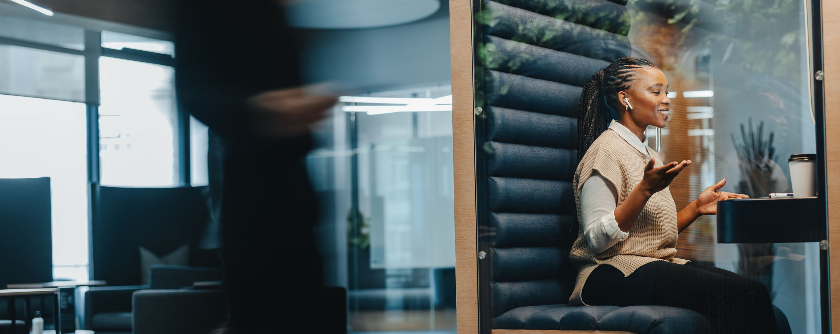 Young businesswoman smiles, sitting in a busy coworking office, discussing and succeeding in her virtual video call with other professionals. Female professional doing remote work from a booth.