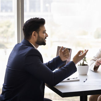 Positive married couple consulting financial expert, real estate agent at meeting, listening to young Indian business consultant in formal suit, sitting at table with paper agreement