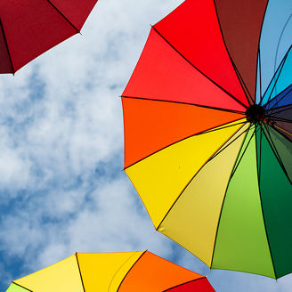 Das Bild zeigt mehrere bunte Regenschirme, die in einem Regenbogenmuster gestaltet sind, vor einem klaren blauen Himmel mit weißen Wolken. Die Schirme sind am oberen Bildrand positioniert und ihre lebendigen Farben kontrastieren stark mit dem blauen Himmel und den fluffigen Wolken, wodurch ein fröhliches und lebendiges Bild entsteht.