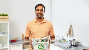 metal recycling, waste sorting and sustainability concept - smiling young indian man in striped t-shirt holding plastic box with tin cans over home kitchen background