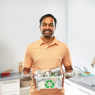 metal recycling, waste sorting and sustainability concept - smiling young indian man in striped t-shirt holding plastic box with tin cans over home kitchen background