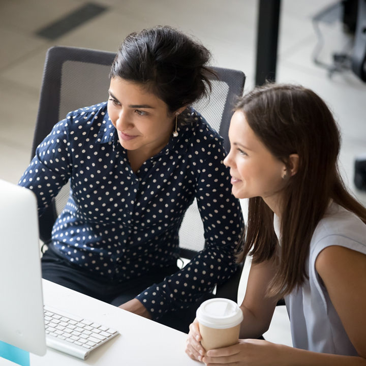 Multiracial colleagues indian and caucasian young women having coffee break sitting together at desk in office. Diverse students interns friends looks at pc screen talking discussing working moments