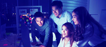 Four people gathered around a desktop computer, leaning toward the screen, with a keyboard, mouse, and office items on the desk in a dimly lit workspace