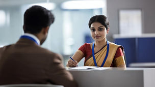 Indian woman sitting at a bank counter and a man in a suit sitting behind her, discussing or offering incentives, financial consultation, bank transaction assistance