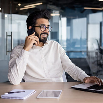 A smiling young Indian man is sitting at a desk in the office, using a laptop and talking on the phone.