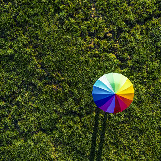 Das Bild zeigt eine Luftaufnahme eines bunten Regenschirms, der auf einer grünen Wiese liegt. Der Schirm ist in einem Regenbogenmuster gestaltet, mit Farben von Rot über Orange, Gelb, Grün, Blau bis hin zu Violett. Die Wiese ist dicht mit Gras bewachsen und bietet einen natürlichen Hintergrund. Der Schirm wirft einen langen Schatten auf das Gras, der durch die tief stehende Sonne entsteht. Der Kontrast zwischen den lebendigen Farben des Schirms und dem grünen Gras macht das Bild besonders auffällig.