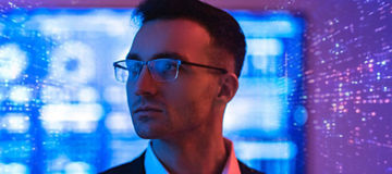 Close-up of a man in a suit wearing glasses, facing slightly to the side, with a blurred background of blue and purple digital lights and abstract patterns