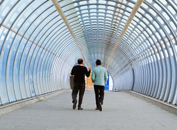 Two individuals walking through a modern enclosed pedestrian walkway with a curved glass and metal structure.