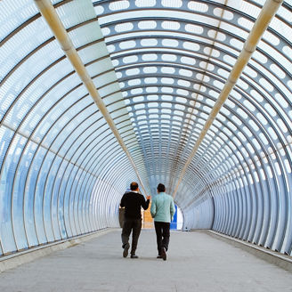 Two individuals walking through a modern enclosed pedestrian walkway with a curved glass and metal structure.